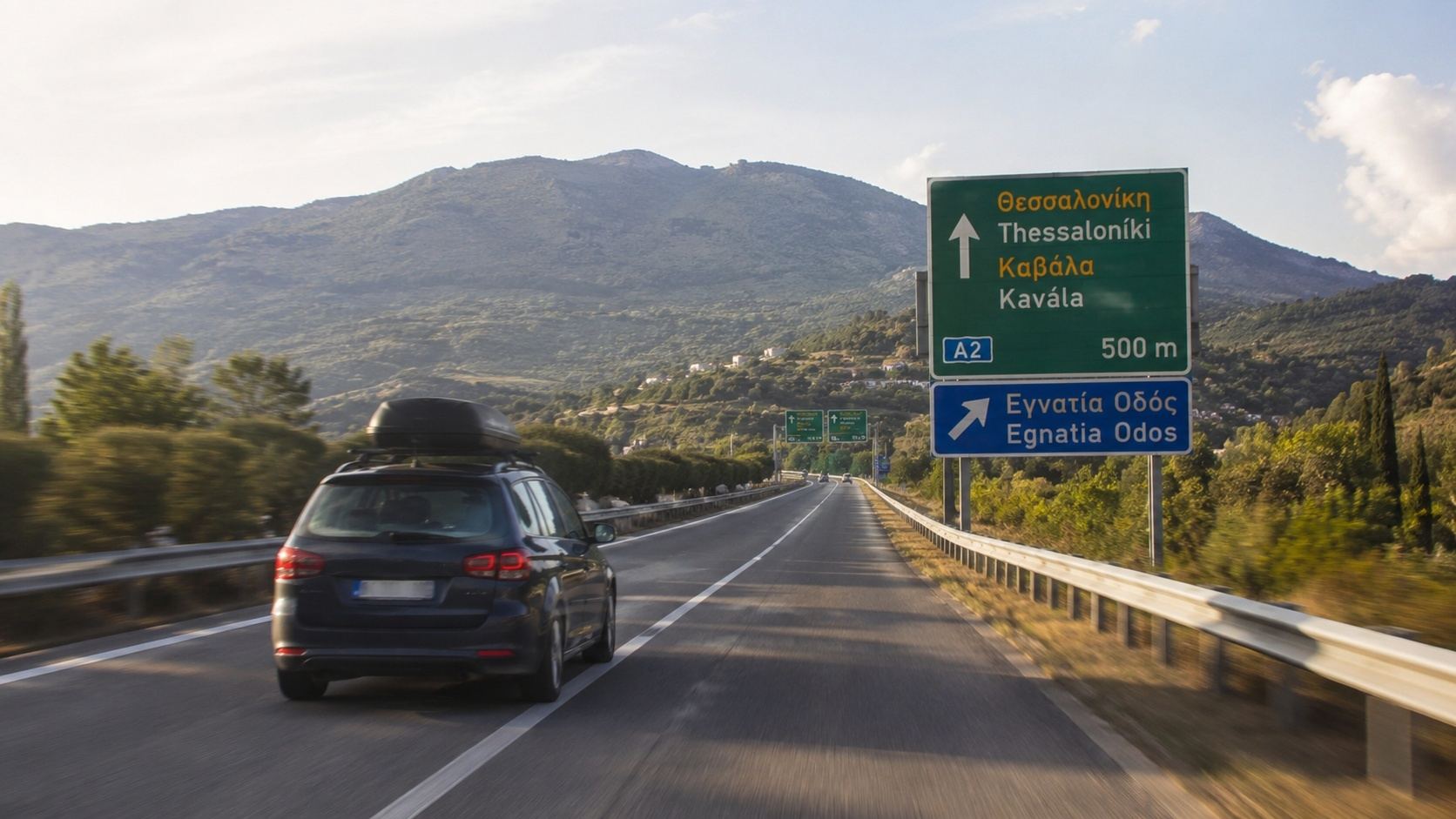 Automobil sa krovnim koferom vozi autoputem kroz Grčku, pored putokaza za Solun i Kavalu.