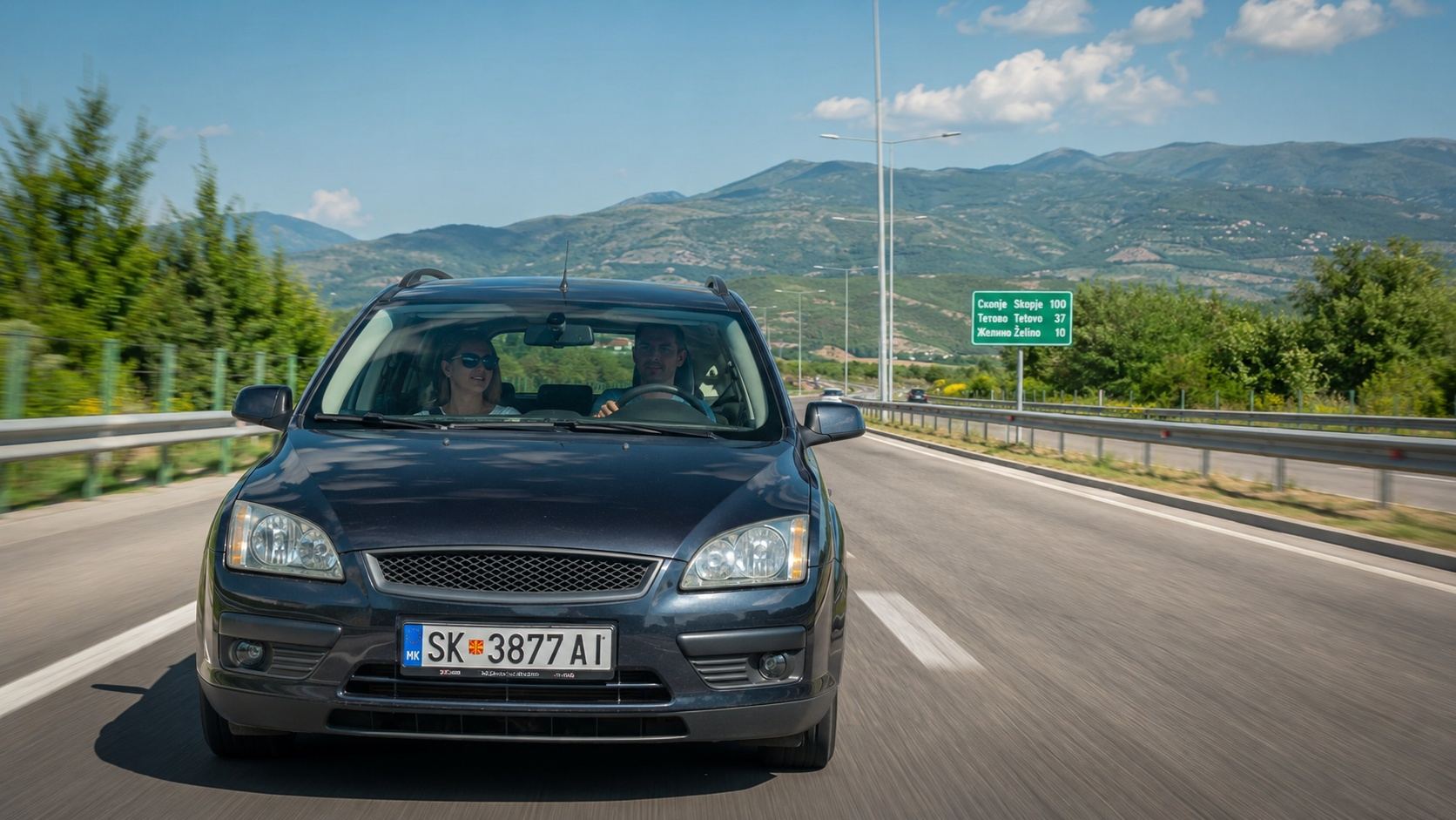 Muškarac i žena putuju automobilom autoputem kroz Severnu Makedoniju, uz planinski pejzaž i putokaz ka Skoplju.